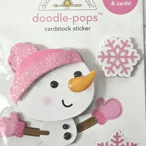 May include: Cardstock sticker set featuring a snowman with a pink hat, mittens, and a carrot nose, along with two pink and white snowflakes. The sticker set is labeled "doodle-pops" and is designed for scrapbooks and cards.