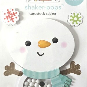 May include: A cardstock sticker featuring a snowman design with a shaker element. The snowman has a carrot nose, black eyes, and a blue scarf. The package includes snowflake stickers and text that reads "shaker-pops" and "perfect for scrapbooks & cards!"