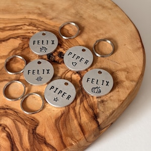 May include: A selection of silver pet tags, each stamped with a name and a symbol such as a paw print, leaf, heart, star, or pumpkin. Small metal rings are included. The tags are displayed on a wooden board.