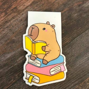 May include: A whimsical bookmark featuring a cartoon capybara reading a yellow book while seated atop a stack of colorful books. The capybara is light brown with a pink cheek. The books are blue, pink, and yellow.