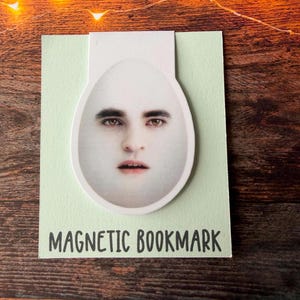 May include: A magnetic bookmark featuring an egg-shaped cutout with a printed human face. The bookmark is attached to a light green rectangular card with the text MAGNETIC BOOKMARK printed in black at the bottom of the card surface.