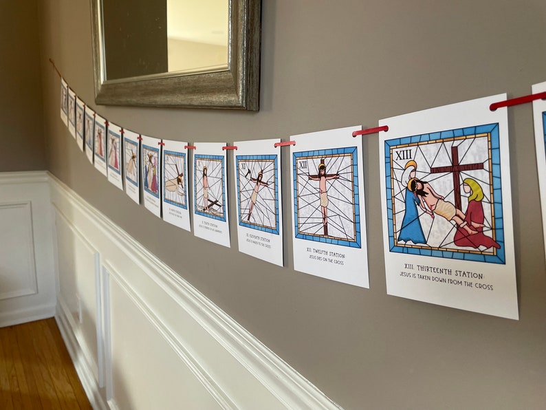 May include: A white wall with a string of 13 cards hanging from it. Each card features a stained glass window design depicting a scene from the Stations of the Cross. The cards are numbered 1 through 13 and each card has a short description of the scene. The cards are held up by red clothes pegs.