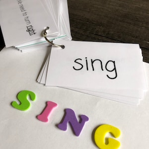 May include: A set of white flashcards with the word "sing" written on them. The word "sing" is spelled out with colourful foam letters.