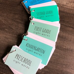 May include: A set of five colourful flashcard sets for different grade levels, starting with preschool and ending with third grade. Each set has a different colour and includes the words "sight words" on the front.