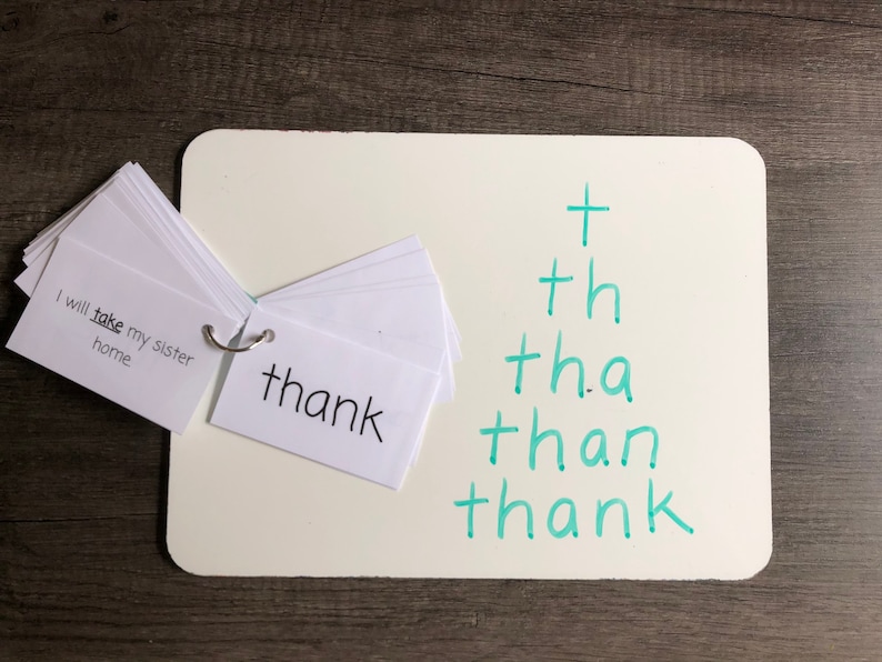 May include: A white board with the word "thank" written on a white card and a green phonics chart with the words "t", "th", "tha", "than", and "thank" written on it.