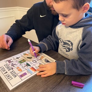 May include: A father and son are playing a Lenten Bingo game. The game board features images related to the Christian season of Lent, such as a cross, a crown of thorns, and a dove. The boy is using a purple marker to mark off a square on the board.