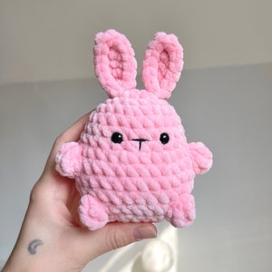 May include: A handmade pink crochet bunny with black embroidered eyes and a small black stitched mouth. The bunny has long ears and small arms. The plush toy is made of soft, textured yarn.