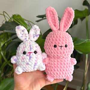 Puede incluir: Dos peluches de conejo de crochet. Uno es rosa y el otro es blanco con detalles rosas y morados.