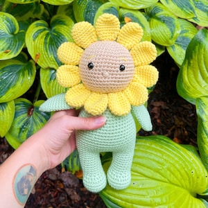 Pode incluir: Um brinquedo de girassol de crochê com um corpo verde e pétalas amarelas. O brinquedo tem um rosto marrom com olhos pretos e um pequeno sorriso costurado.