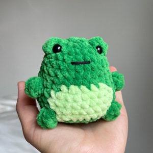 May include: A green crocheted frog plush toy with black eyes and a black stitched mouth. The frog has a light green belly.