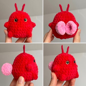 May include: A red crocheted plush toy with pink accents. The toy has a round body with two small antennas on top. The toy has two large eyes and a small mouth.