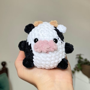 Puede incluir: Un juguete de peluche de vaca tejido a crochet con pelaje blanco y negro, orejas marrones y nariz rosa.