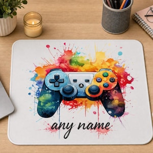 May include: A rectangular mouse pad featuring a central graphic of a video game controller surrounded by a vibrant, multi-coloured watercolour paint splatter effect. The text "any name" is printed in a black cursive font below the controller.