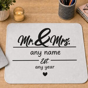 May include: A rectangular white mouse pad featuring black text that reads Mr. & Mrs. followed by any name, Est. any year, and a small black heart icon centred at the bottom. The design is printed on a smooth surface with rounded corners.