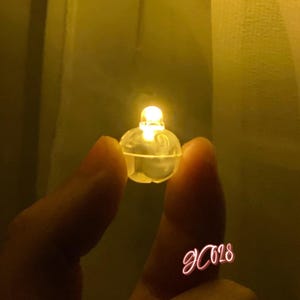 May include: A small, spherical, translucent plastic LED light emitting a warm yellow glow. The device features a rounded top section with a clear bulb and a slightly wider base, creating a compact, illuminated component for decorative lighting.