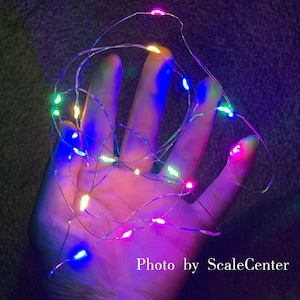 Op de afbeelding: Een hand die een reeks veelkleurige LED-lampjes vasthoudt. De lampjes zijn klein en rond, met kleuren als roze, blauw, groen en geel. De lampjes zitten op een dunne, zilverkleurige draad. De tekst "Photo by ScaleCenter" staat in de rechter benedenhoek.