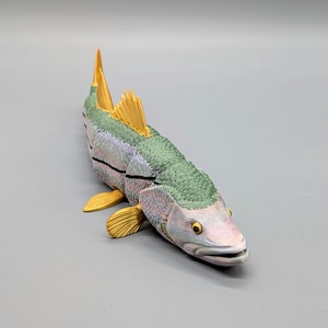May include: A multi-jointed fish figurine featuring a textured green back, a pale pinkish-white underbelly, and bright yellow fins. The body is segmented with visible black lines between sections, and the head has a realistic eye and open mouth.