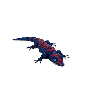 Könnte beinhalten: Ein Stofftier in Form einer Eidechse. Der Körper ist dunkelblau mit roten Markierungen. Die Eidechse hat vier Beine, einen langen Schwanz und blaue Augen. Das Spielzeug ist als Gecko gestaltet.