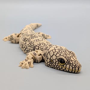 Peut inclure: Une figurine de lézard articulée présentant un corps de couleur crème avec un motif d'écailles brun foncé détaillé sur le dos, la tête et les membres. Le reptile possède une structure corporelle segmentée, des articulations visibles et des yeux sombres.