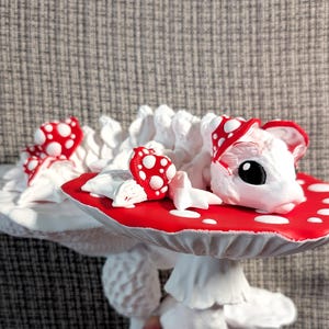 Könnte beinhalten: Eine skurrile Pilzskulptur mit einer rot-weiß gepunkteten Kappe. Eine weiße Maus mit einem rot-weiß gepunkteten Hut ruht auf dem Pilz. Die Skulptur hat einen weißen Stiel und ein strukturiertes, geschichtetes Design.