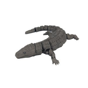 Op de afbeelding: Een grijze, gelede alligatorfiguur met een getextureerd oppervlak. De speelgoedalligator heeft een gesegmenteerd lichaam, vier kleine poten en een gedetailleerde kop. De alligator is neutraal grijs van kleur en staat tegen een witte achtergrond.