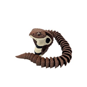 Könnte beinhalten: Braune und beige Cobra-Schlangenfigur mit detailliertem Kopf und segmentiertem Körper. Der Kopf der Schlange ist erhoben und zeigt ein schwarz-weißes Muster auf dem Hals. Die Figur besteht aus einem festen Material.