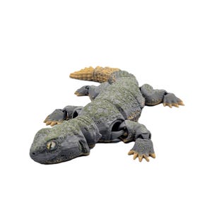 May include: A detailed, articulated lizard figurine in shades of grey, green, and tan. The reptile has a textured body, visible joints, and detailed claws. The figurine is posed on a white background, showcasing its realistic design.