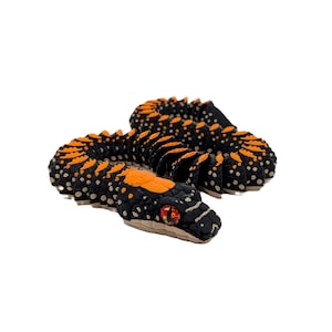 Puede incluir: Una serpiente de juguete negra y naranja. La serpiente presenta un diseño detallado con marcas naranjas sobre un cuerpo negro. La cabeza tiene ojos rojos. El juguete probablemente está hecho de un material blando.