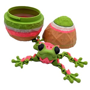 Könnte beinhalten: Ein grünes und pinkfarbenes Froschspielzeug mit beweglichen Gliedmaßen aus Kunststoff. Der Frosch ist mit ausgestreckten Beinen in Pose. Der Frosch sitzt auf einem grün-braunen eiförmigen Behälter mit Deckel.