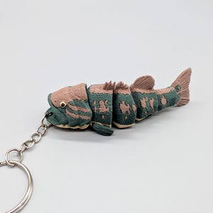 Peut inclure: Un porte-clés poisson articulé présentant un corps texturé avec une palette de couleurs vert sourd et rose poudré. La figurine de poisson segmentée est fixée à un anneau fendu en métal argenté par une courte chaîne pour plus de souplesse.