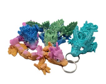Pode incluir: Uma coleção de chaveiros de dragão coloridos em tons de azul, verde, rosa e laranja. Cada dragão tem uma cabeça detalhada e um corpo segmentado, preso a um chaveiro prateado. Os chaveiros estão dispostos sobre um fundo branco.