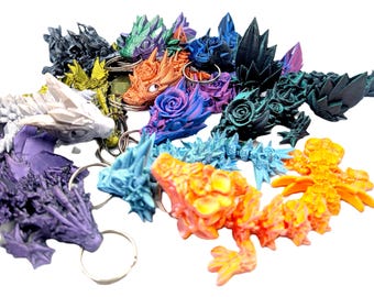 Pode incluir: Uma coleção de porta-chaves de dragão articulados em várias cores, incluindo laranja, azul, roxo, branco, preto e verde. Cada dragão apresenta texturas complexas de escamas e rosas na cabeça e no corpo, presos a um anel de metal.