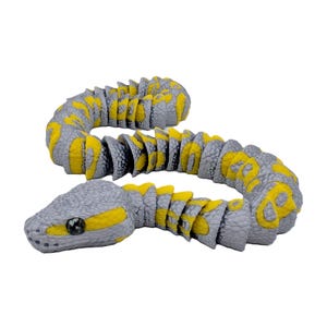 Op de afbeelding: Een grijze en gele speelslang met een gesegmenteerd lichaam. Het lichaam van de slang is grijs met gele markeringen en heeft een grijze kop met een zwart oog. Het speelgoed is ontworpen om op een echte slang te lijken.
