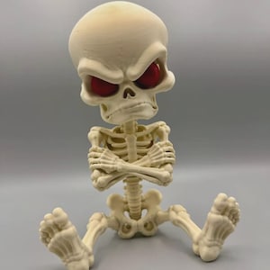 May include: A cream-colored skeleton figurine with a large, stylized head and deep-set, solid red eyes. The skeleton is depicted in a seated position with its arms crossed over its chest, featuring a prominent brow ridge and a neutral, bony texture.