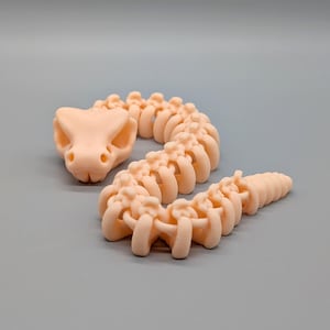 Pode incluir: Um brinquedo de esqueleto de cobra articulado, cor de pêssego. A cabeça da cobra tem órbitas oculares e um focinho. A coluna vertebral é feita de vértebras conectadas, e a cauda afunila-se até um ponto.