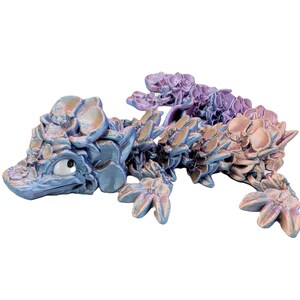 May include: A whimsical, articulated dragon figurine in shades of blue, purple, and peach. The dragon is crafted with a floral-like texture, giving it a unique, fantasy aesthetic. The dragon has a single eye and is posed on a white background.