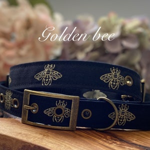 May include: Black dog collar with a gold metal buckle and gold bee design. The collar is made of fabric and has multiple eyelets for adjusting the size.
