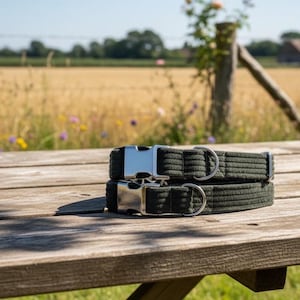 Könnte beinhalten: Zwei olivgrüne Hundehalsbänder mit silbernen Metallschnallen und D-Ringen sind auf einem hölzernen Picknicktisch gestapelt. Die Halsbänder haben eine gerippte Textur. Der Hintergrund zeigt ein Weizenfeld und einen blauen Himmel.