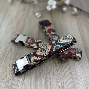 May include: Two pet collars featuring a woven fabric strap with a multi-coloured geometric pattern in shades of blue, red, yellow, and cream. Each collar includes a silver-toned metal D-ring and a black plastic side-release buckle fastener.