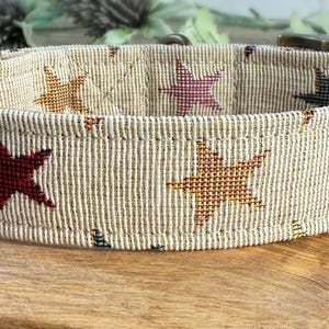 Pu&ograve; includere: Un collare per cani beige con un motivo a stelle colorate intrecciato. Il collare ha una fibbia in ottone e un anello a D in ottone.