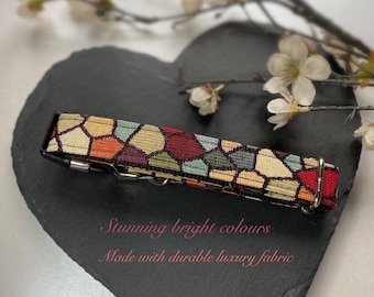 May include: A patterned dog collar featuring a geometric design in shades of red, gold, green, and orange. It has a metal buckle and a black backing. The image text says "Stunning bright colours" and "Made with durable luxury fabric."