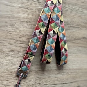 May include: A colorful dog leash with a geometric pattern of triangles in red, green, blue, and yellow. The leash has a silver metal clasp and a black underside. The leash is made of a woven fabric.