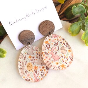 May include: A pair of wooden and resin earrings with a floral and bunny design. The earrings are oval shaped and feature a brown wooden disc at the top. The text "Wandering Woods Designs" is visible on a white card behind the earrings.