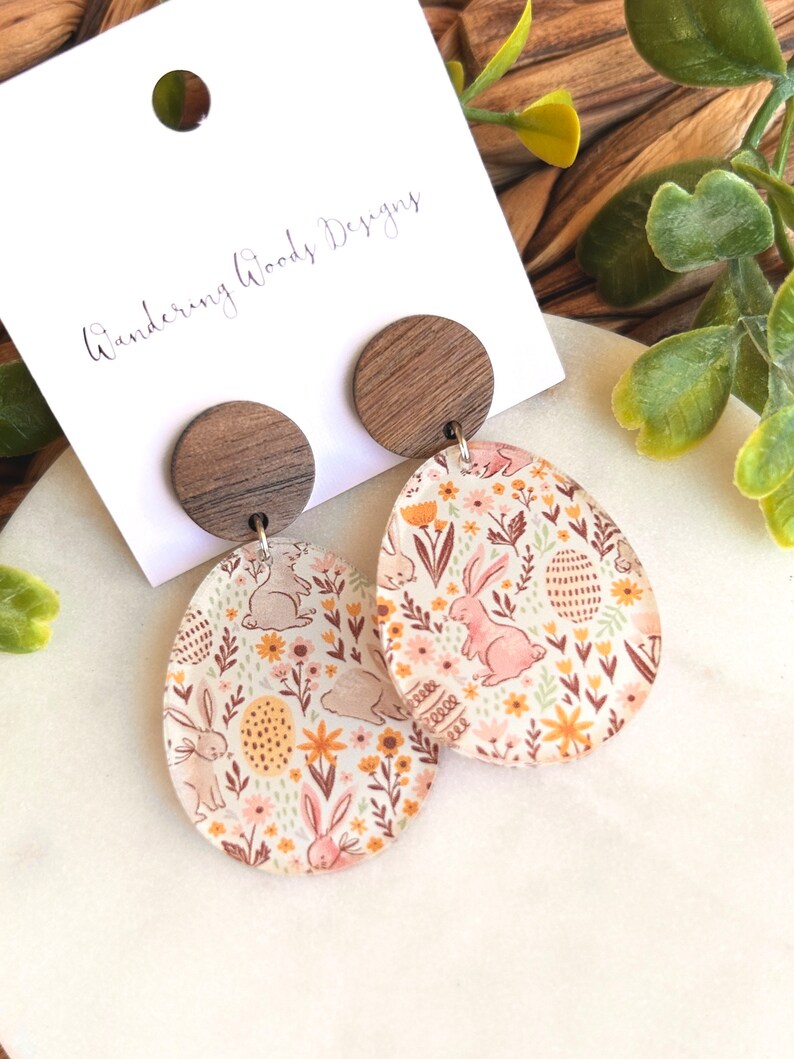 May include: A pair of wooden and resin earrings with a floral and bunny design. The earrings are oval shaped and feature a brown wooden disc at the top. The text "Wandering Woods Designs" is visible on a white card behind the earrings.