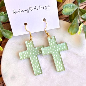 May include: A pair of green cross earrings with a white polka dot pattern. The earrings are made of acrylic and have gold hooks.