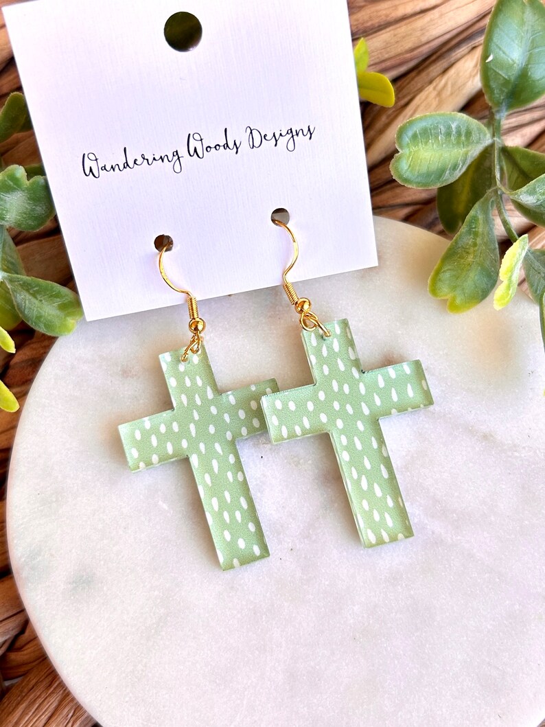 May include: A pair of green cross earrings with a white polka dot pattern. The earrings are made of acrylic and have gold hooks.