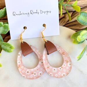 May include: A pair of pink floral dangle earrings with brown leather accents. The earrings are on a white surface with a card that says "Wandering Woods Designs".