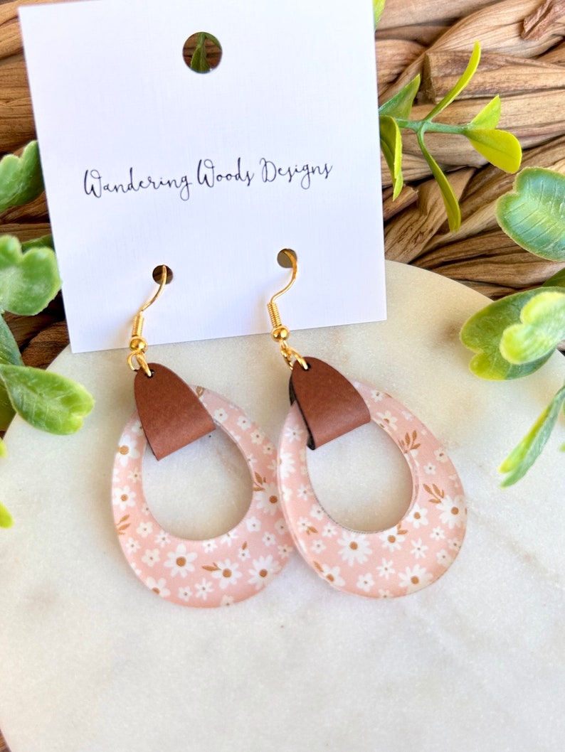 May include: A pair of pink floral dangle earrings with brown leather accents. The earrings are on a white surface with a card that says "Wandering Woods Designs".