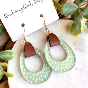 May include: A pair of green and white polka dot earrings with brown leather accents. The earrings are in the shape of a circle with a cutout in the middle. The earrings are hanging from gold hooks.