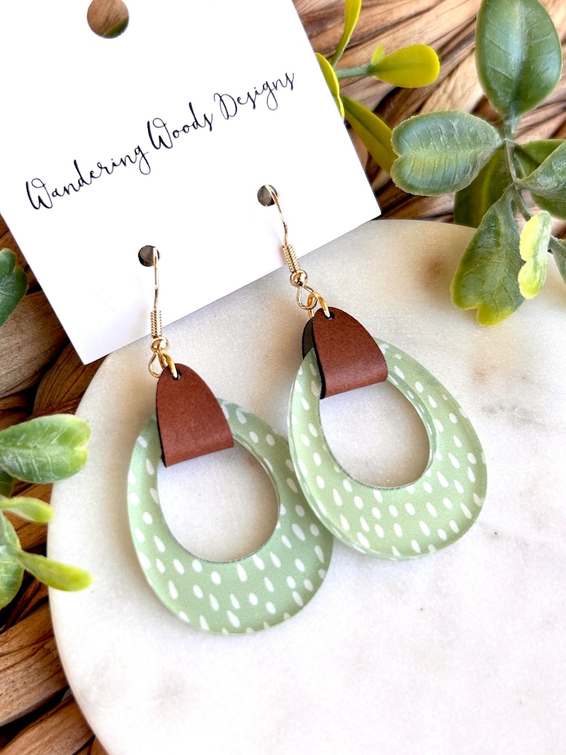 May include: A pair of green and white polka dot earrings with brown leather accents. The earrings are in the shape of a circle with a cutout in the middle. The earrings are hanging from gold hooks.
